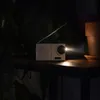 A small, silver portable radio with an antenna is placed on a wooden shelf in a dimly lit room. The radio is emitting a bright light from its speaker, illuminating the surrounding area. There are dark green plants and a brown terracotta pot in the backgrou Voir les images du produit