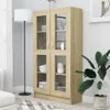 A tall, light brown wooden display cabinet with glass doors is shown in a room with a floor lamp and a potted plant. Produktbild-Vorschau