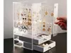 A hand is shown opening a drawer of a clear acrylic jewelry organizer box filled with various necklaces and earrings. Produktbild-Vorschau