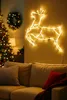 A living room decorated for Christmas with a lit Christmas tree, a wreath, and a reindeer-shaped light decoration on the wall above a sofa. Produktbild-Vorschau