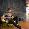 A man is playing an electric guitar while sitting on a beanbag chair in a room with a brick wall. Náhľad obrázku produktu