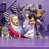 Two young girls are playing with LEGO race cars and track pieces on a purple table, with a purple workshop-themed background. Produktbild-Vorschau