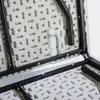A close-up view of the underside of a white folding table, showing the metal frame and plastic surface with its distinctive hole pattern. A silver L-shaped tool is attached to the plastic surface. Voir les images du produit
