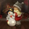 A decorative figurine of a girl hugging a snowman in front of a fireplace. Produktbild-Vorschau