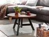 A round wooden coffee table with a black metal frame, placed in a living room setting next to a grey sectional sofa. A plant, a pastry, and a cup of coffee are on the table. A wooden crate with a camera is on the floor. Náhľad obrázku produktu