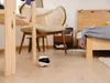 A small robot is on a wooden floor in a bedroom next to a chair and a bed. Náhled obrázku produktu
