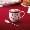 A Christmas-themed mug filled with hot chocolate sits on a red tablecloth next to a spoon. A cake is visible in the background. Náhled obrázku produktu