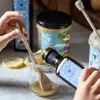A person is pouring oil from a bottle into a glass jar with a wooden spoon and lemon slices nearby. Produktbild-Vorschau
