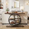 A console table with a circular shelf unit is placed behind a sofa in a living room setting. The table has a wooden top and shelves, with a black metal frame. Decorative items like books, a bust, and a vase are placed on the shelves. Anteprima dell’immagine prodotto