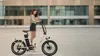 A young woman stands next to a cream-colored electric bicycle on a paved outdoor plaza in front of a modern building. Produktbild-Vorschau