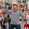 A man dressed as a sailor with a spyglass is in the foreground of a carnival parade with confetti falling. Produktbild-Vorschau