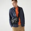 A man wearing a blue shirt and orange polo shirt, with a black messenger bag, standing against a white background. Anteprima dell’immagine prodotto