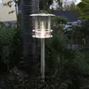 A solar-powered path light is illuminated in a garden at dusk, next to a tree trunk and surrounded by plants. Produktbild-Vorschau