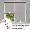 A window with a roller blind partially lowered, showing a garden view. A hand is shown operating a crank mechanism for the blind. Náhled obrázku produktu