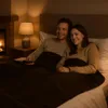 A couple is lying in bed, covered by a blanket, watching TV by the fireplace. Produktbild-Vorschau