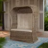 A woven rattan chair with cushions sits on a rug on a wooden deck, with doors and foliage in the background. Podgląd zdjęcia produktu