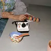 A person's hands are shown on a table next to a Polaroid camera, a bowl of ice cream, and a toy. Voir les images du produit