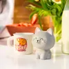 A cute gray cat figurine sits next to a mug with a cookie design, with flowers and strawberries in the background. Voir les images du produit