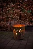A rusty metal fire basket with flames cut into its sides, lit with candles, sits on a brick patio in front of a dense bush with brown leaves. Produktbild-Vorschau