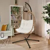A hanging chair with a white cushion is placed in a living room setting, next to a bookshelf and a sofa. Produktbild-Vorschau