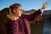 A woman wearing a burgundy sweater stands on a bridge with a river and buildings in the background. Produktbild-Vorschau