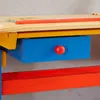 A close-up view of a colorful children's desk, showing a wooden tabletop, a yellow support beam, a blue drawer with a red knob, and a red support beam below. Produktbild-Vorschau