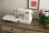 A white sewing machine is placed on a wooden desk, with sewing accessories and a basket of fabric in the background. Produktbild-Vorschau