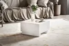 A modern living room scene with a white coffee table in the foreground and a sofa in the background. Produktbild-Vorschau