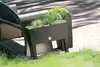 A dark brown planter box filled with various green plants sits next to a dark metal bench on a paved patio with grass in the foreground. Anteprima dell’immagine prodotto