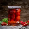 A jar of strawberries in syrup sits on a wooden surface, with loose strawberries and a leaf nearby. Voir les images du produit
