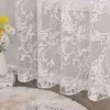 A close-up view of a sheer curtain with an ornate pattern, draped in front of a window. A vase with flowers and books are visible on the left, and a fluffy rug is on the floor in the foreground. Produktbild-Vorschau