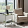 A serving cart with a lamp, cup, and books next to an armchair in a room with a large window. Produktbild-Vorschau