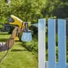 A person is using a yellow and black paint sprayer to paint a light blue fence in a garden setting. Produktbild-Vorschau