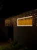 Icicle lights are strung along the roofline of a dark-colored building at night, with a lit window visible. Produktbild-Vorschau