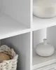 Close-up of white shelving units with decorative items. A basket with coiled rope is in the bottom left. A round, ribbed bowl is on the top right shelf. A small, white vase sits on a stack of books on the bottom right shelf. Náhled obrázku produktu