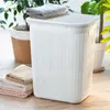 A white laundry basket with a lid sits on a wooden floor, with towels stacked next to it and plants in the background. Voir les images du produit