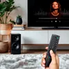 A hand holding a remote control in front of a TV and speaker in a living room. Náhled obrázku produktu
