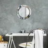 A round mirror with a decorative frame hangs on a textured gray wall above a white desk with a camera and some papers. A chair with a white blanket draped over it is in the foreground. Produktbild-Vorschau