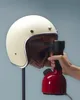 A hand is holding a red and black handheld steamer, spraying steam into a cream-colored helmet that is mounted on a stand. Anteprima dell’immagine prodotto