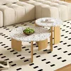 Two round terrazzo coffee tables with wooden legs are placed on a cream-colored rug with a black dashed pattern. The larger table in the foreground holds a bowl of green apples. A portion of a cream-colored sectional sofa is visible in the background. Voir les images du produit
