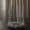 A white candle holder with five white candles arranged in a circle on a dark surface, with a blurred background of vertical blinds. Produktbild-Vorschau