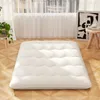 A white mattress is laid out on a wooden floor in front of a window with sheer curtains. Produktbild-Vorschau