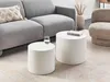Two white cylindrical coffee tables are placed on a rug in a living room setting, next to a gray sofa and a textured armchair. A glass of water sits on the smaller table, and a book rests on the larger one. Náhled obrázku produktu