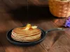 A stack of pancakes with butter and syrup on a small frying pan, placed on a wooden table. Produktbild-Vorschau