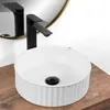A white ceramic washbasin with a black drain and a black faucet is placed on a wooden surface, with a round mirror in the background. Produktbild-Vorschau