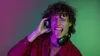 A young man with curly hair wearing headphones and a red shirt, sticking his tongue out and holding one headphone cup. Anteprima dell’immagine prodotto