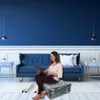 A woman is sitting on a patterned ottoman in a living room, using a laptop. Produktbild-Vorschau