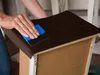 A person is applying dark brown wood-grain contact paper to the top surface of a drawer or cabinet. Produktbild-Vorschau
