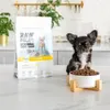 A small dog sits next to a bag of dog food and a bowl of dog food. Produktbild-Vorschau