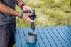 A person is using a water filter to fill a mug on a blue table outdoors. Produktbild-Vorschau